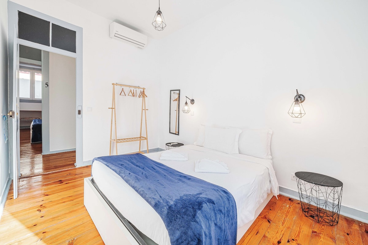 Un dormitorio con una cama, una colcha azul, suelo de madera, un armario y una mesita redonda.