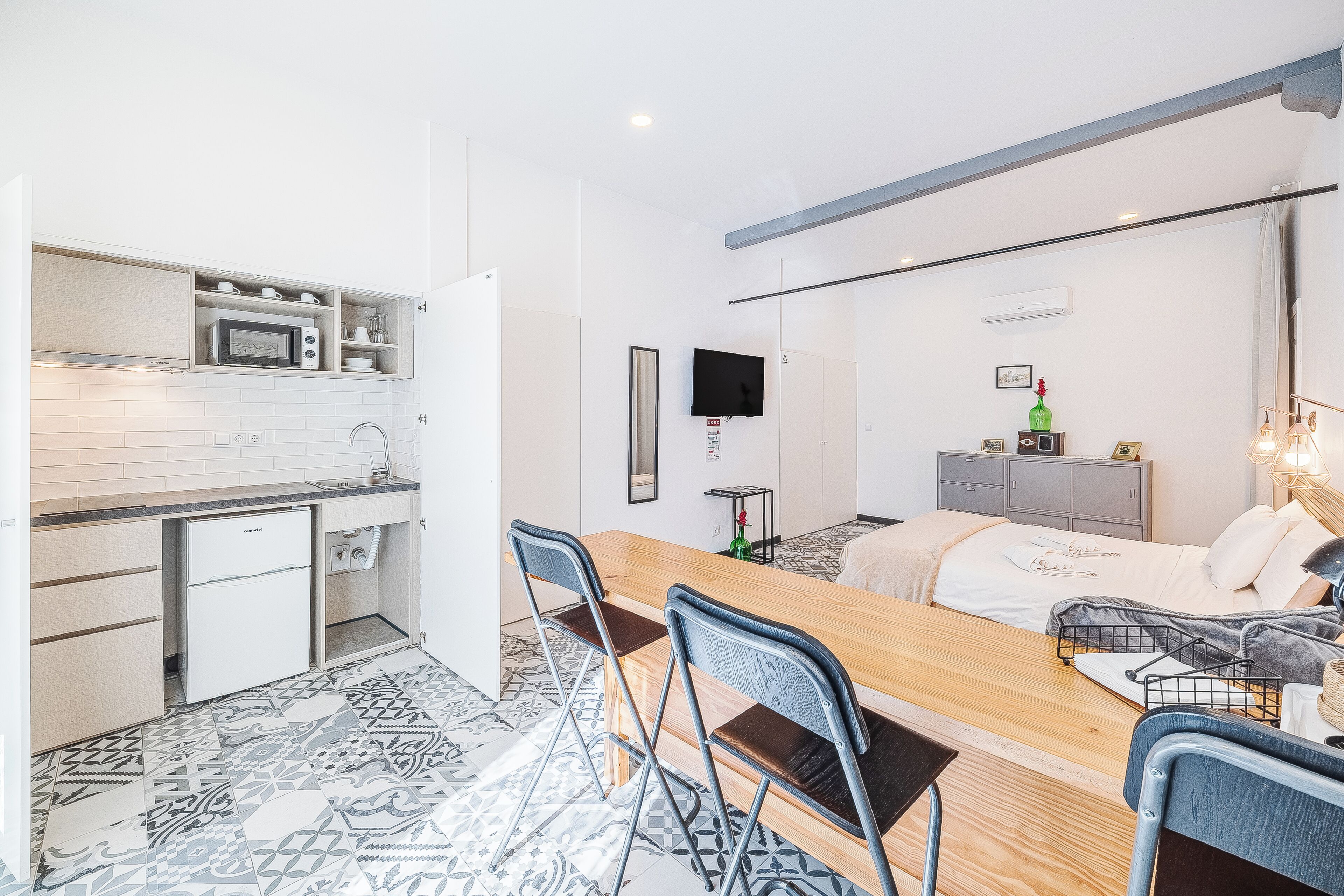 A compact kitchen with a microwave, a small refrigerator, and a sink. There is a dining area with a wooden table and chairs. The room has a bed with a white comforter and a bedside table with a lamp.