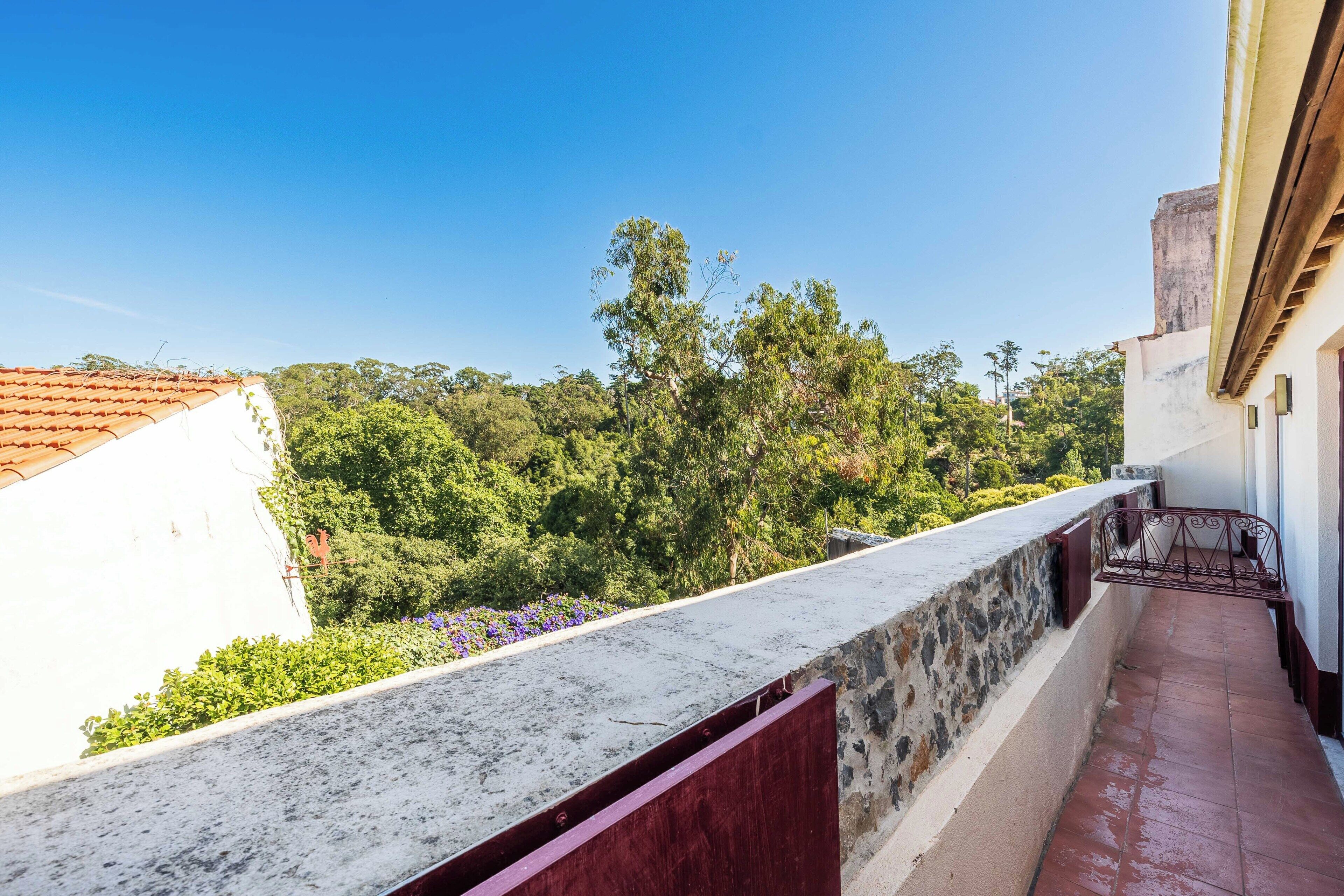 Un balcon doté d’un mur en pierre, d’une rambarde en métal et offrant une vue sur des arbres et un immeuble.