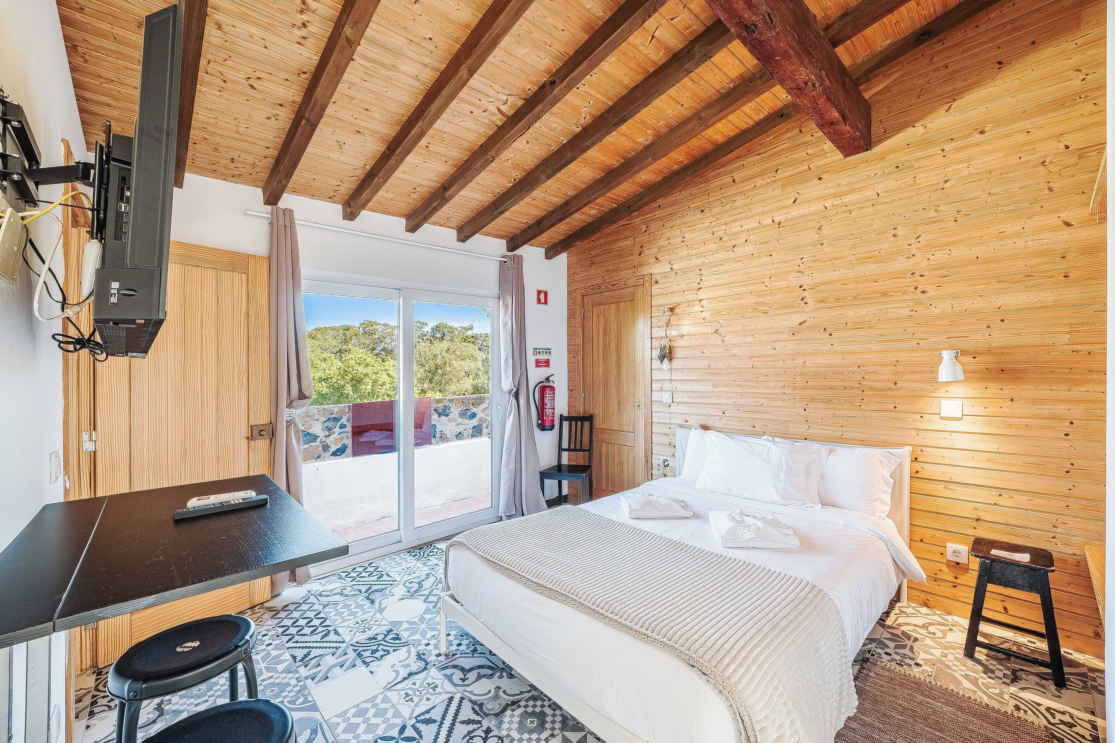 Un dormitorio con cama, escritorio, silla, taburete pequeño, extintor y vistas al exterior.