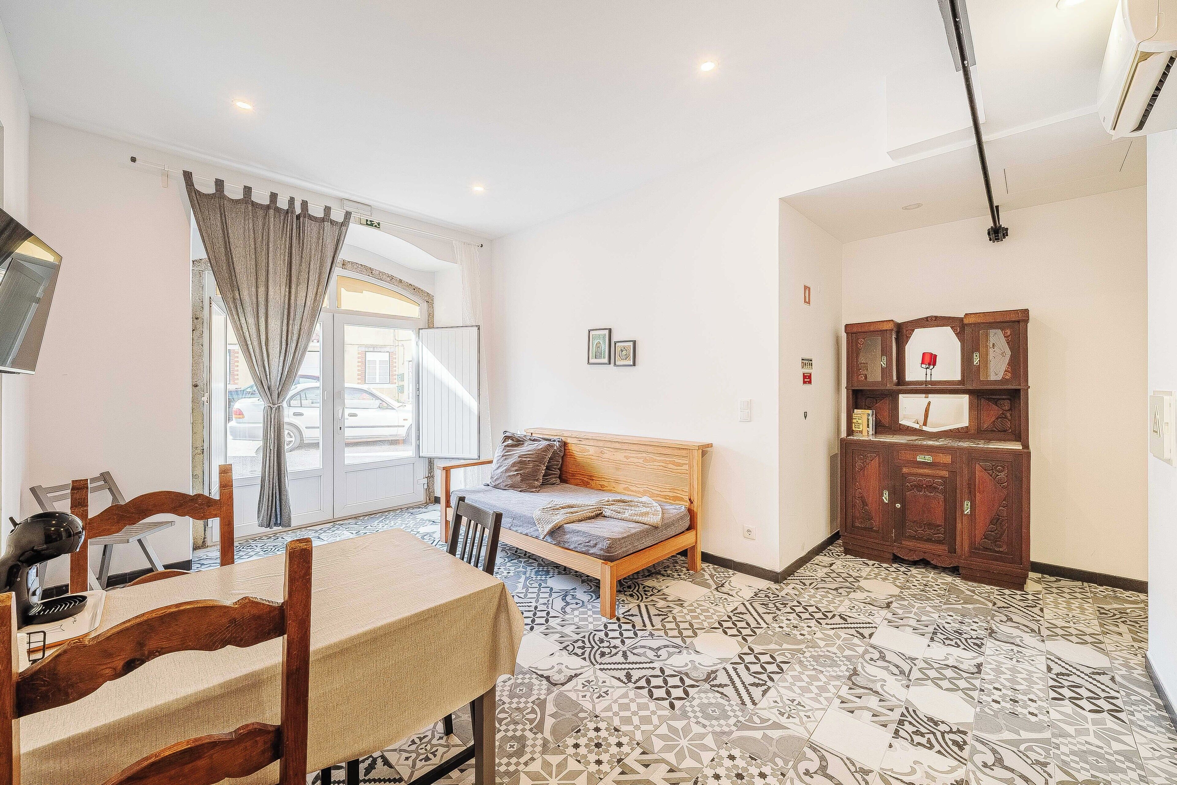 Una habitación con una cama, una mesa de comedor con sillas, piso estampado, un armario y una ventana con cortinas.