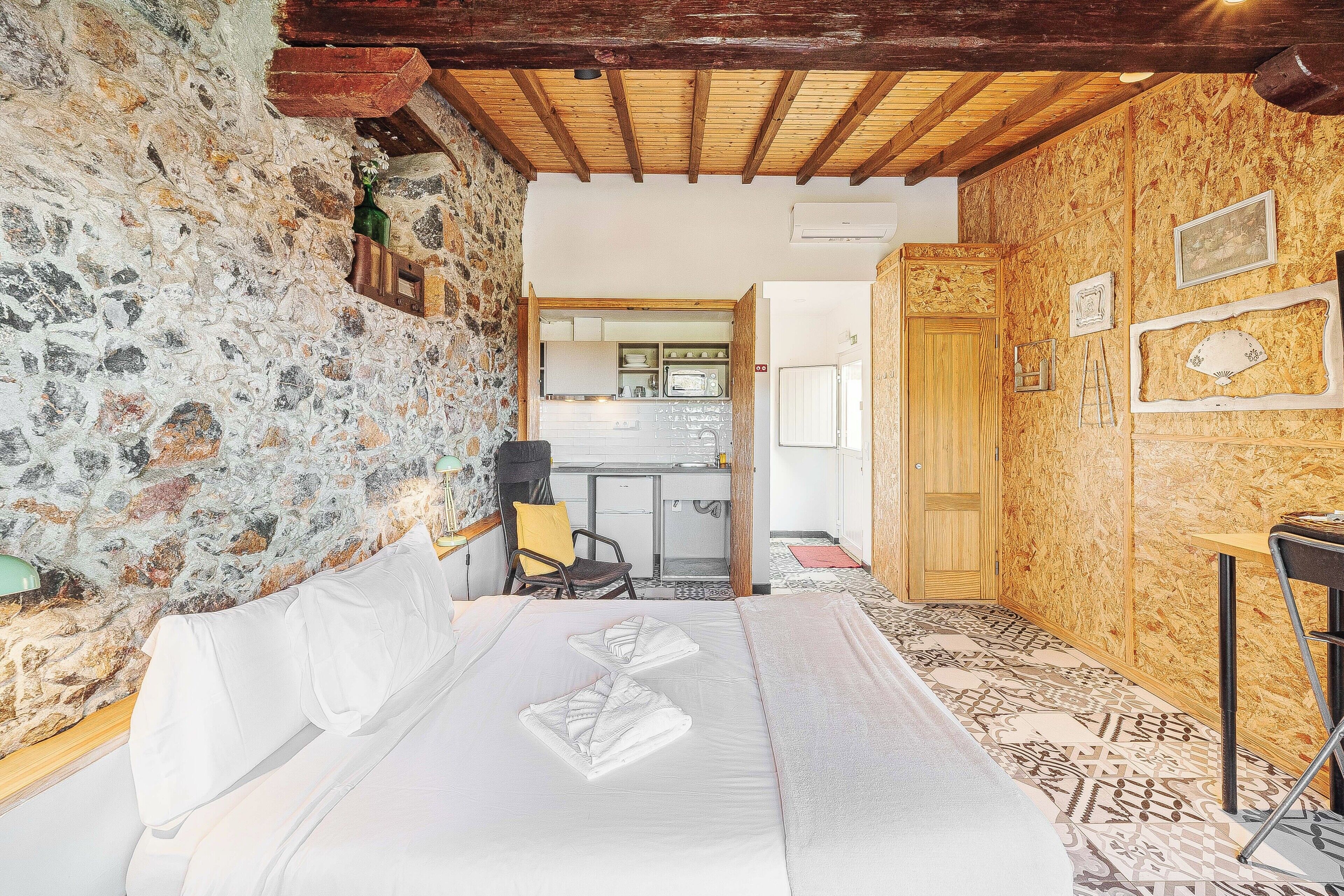 A room with a stone wall, wooden ceiling beams, a bed with white linens, a small kitchenette, and a wooden door.