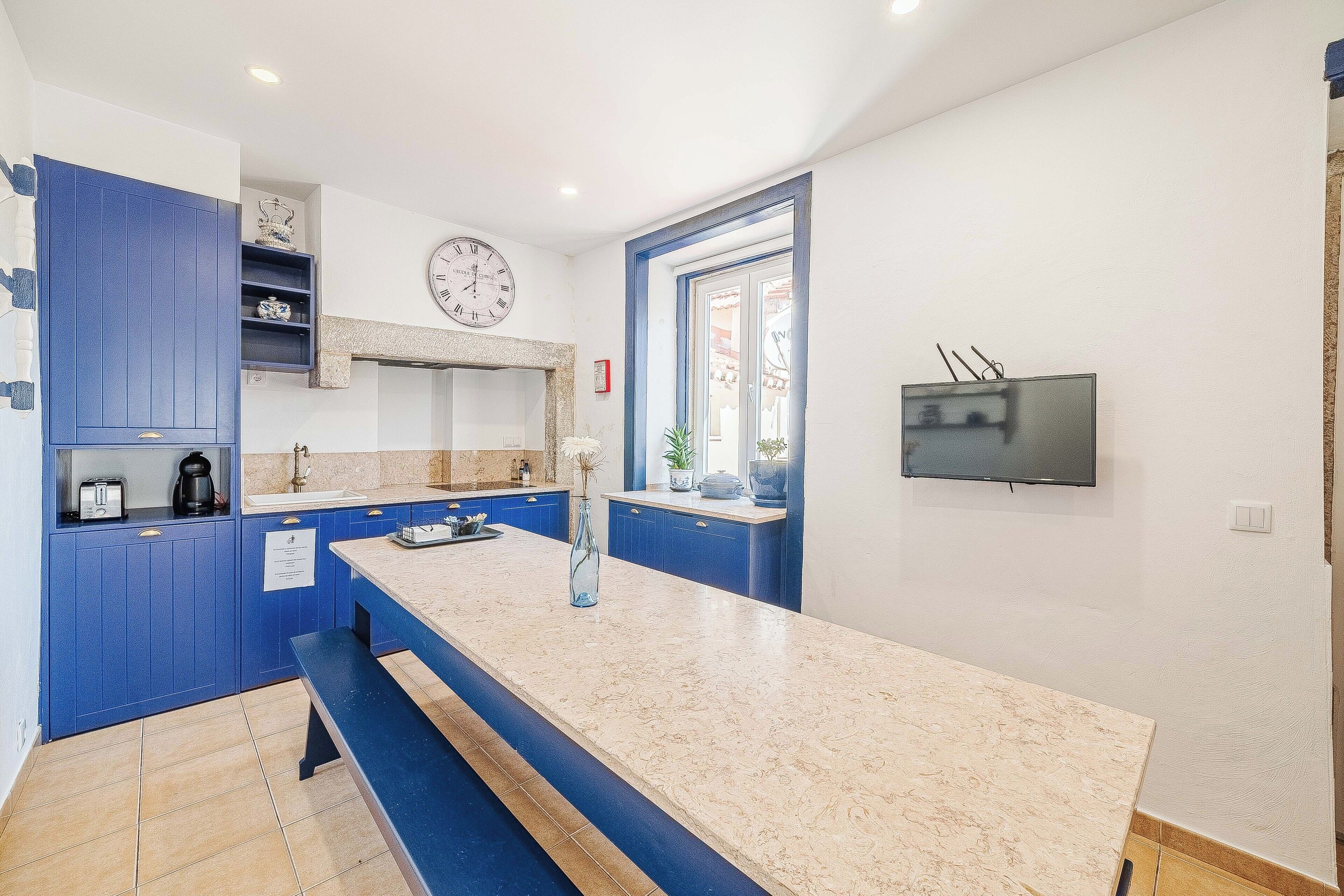 A modern kitchen with blue cabinets, a marble countertop, a built-in oven, and a TV mounted on the wall.