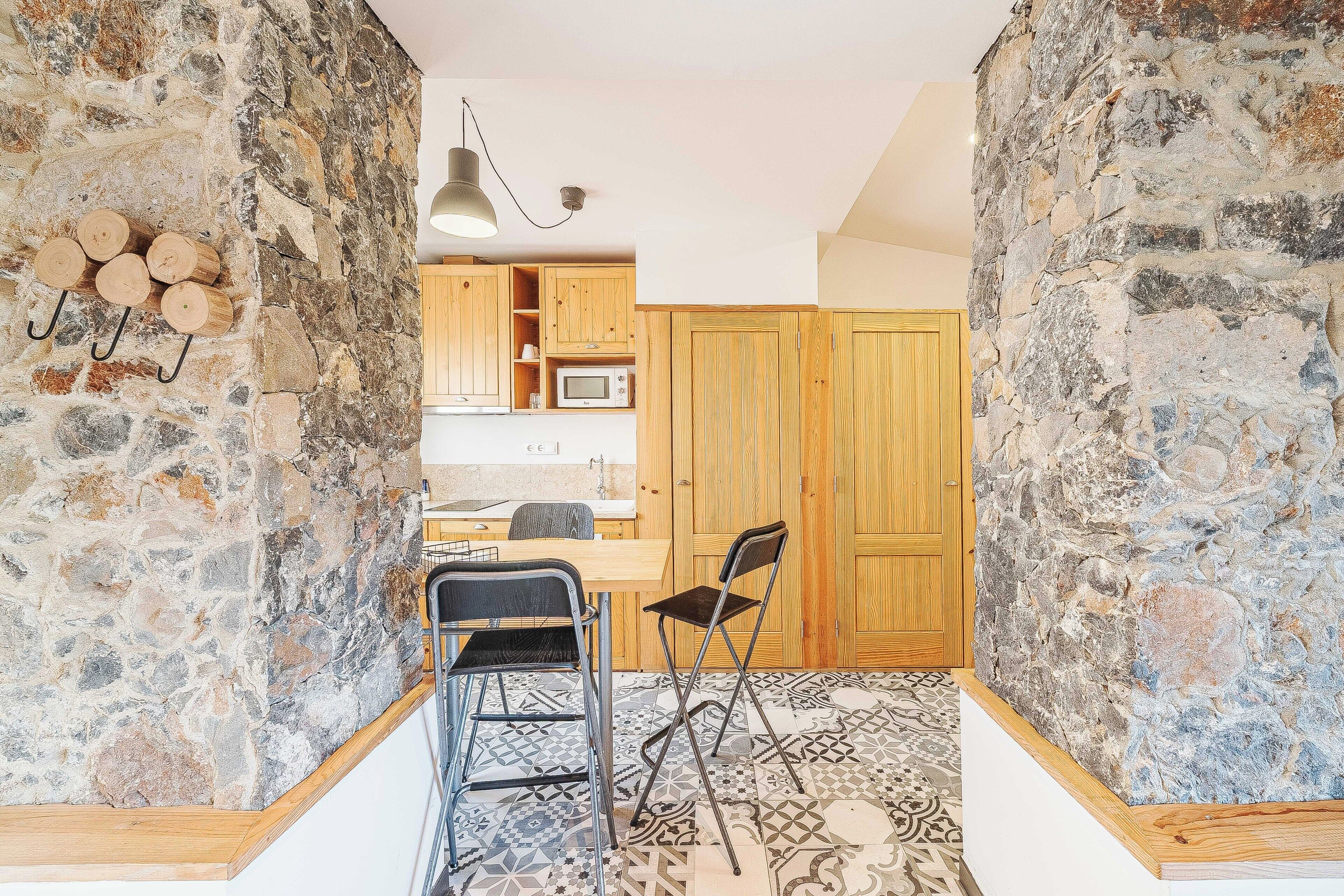 Cocina con paredes de piedra, armarios de madera, una zona de comedor con sillas y suelo con diseño.