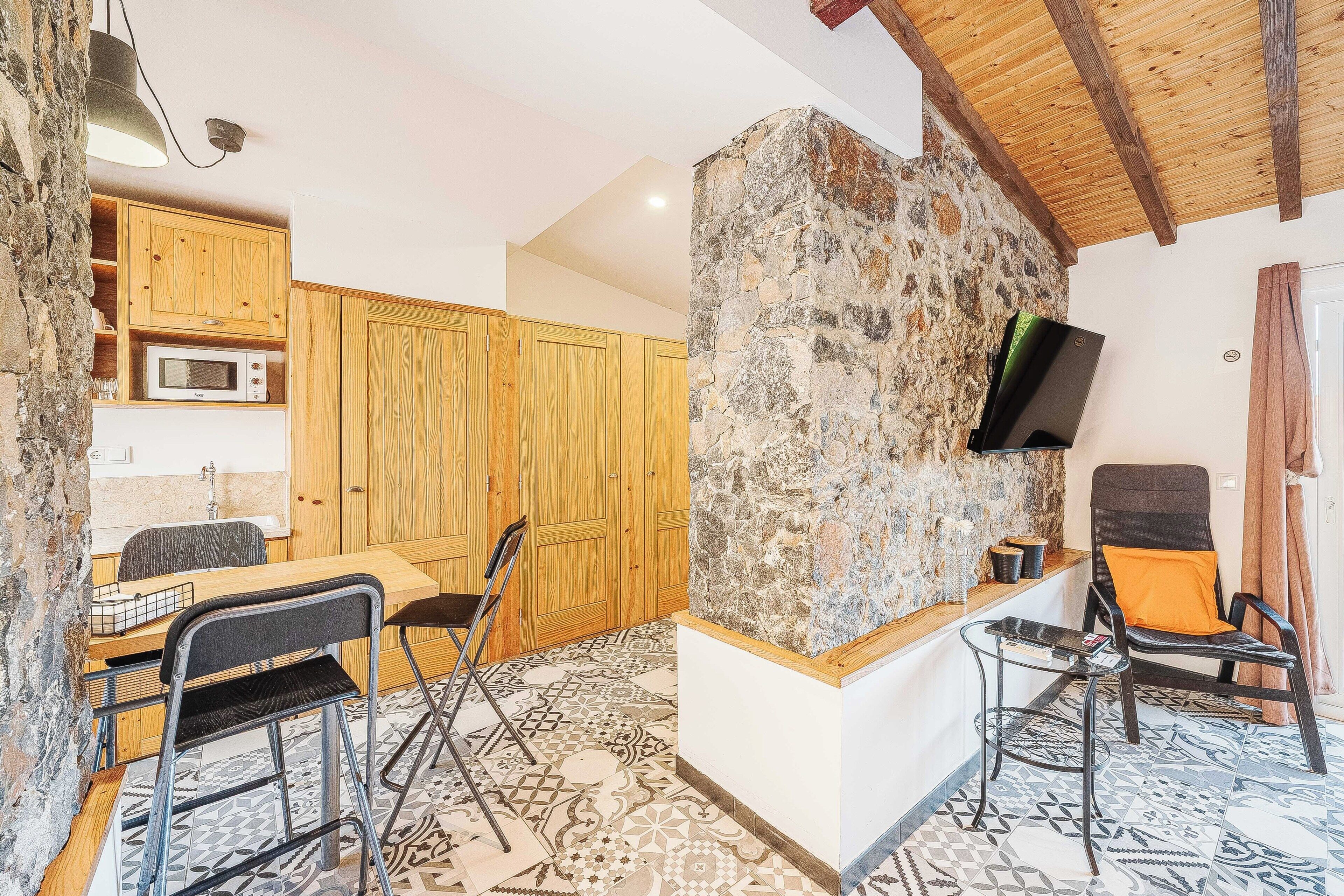 Une cuisine avec des meubles en bois, un mur décoratif en pierre, un téléviseur à écran plat et un coin repas avec des chaises et une table.