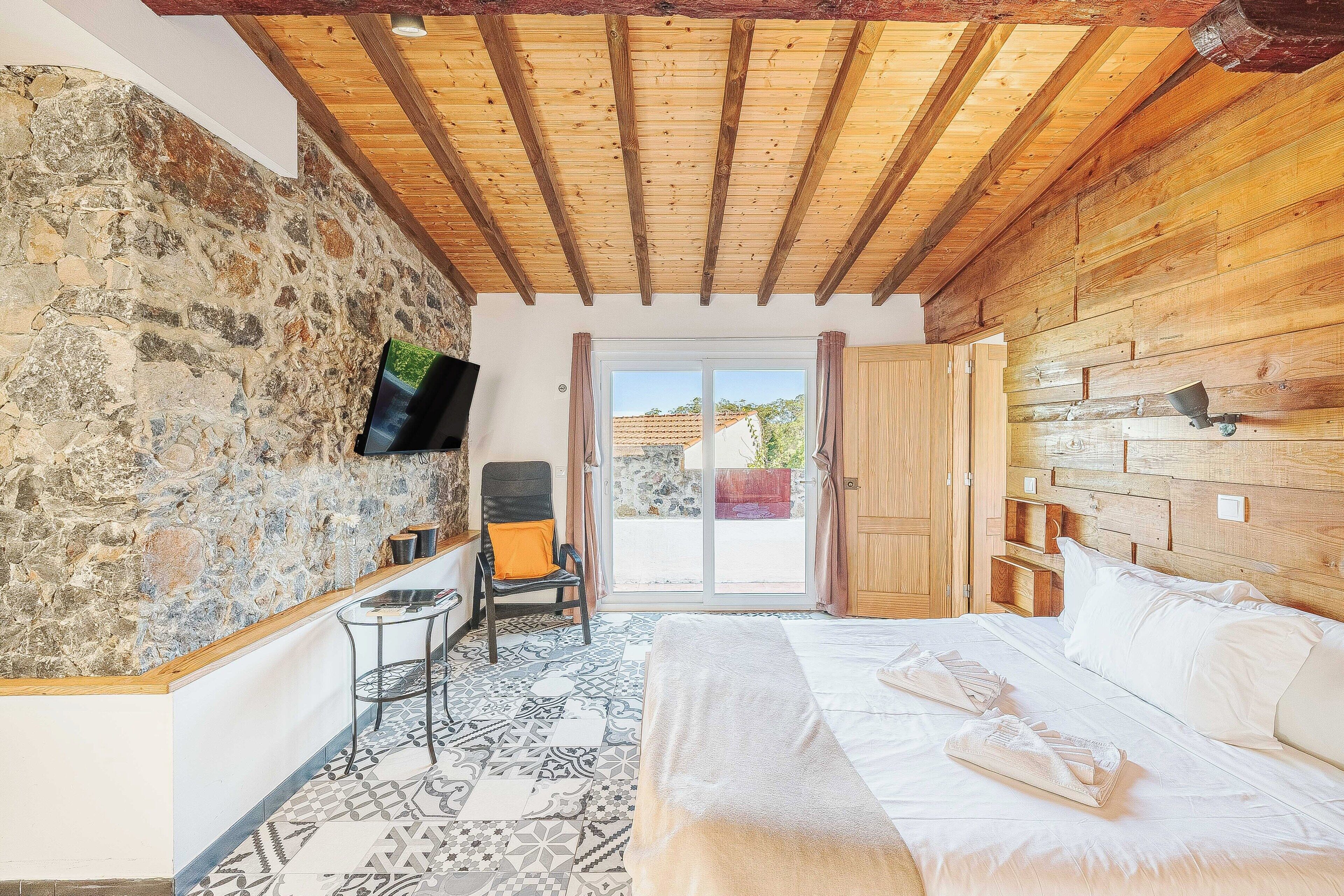 A room with a stone wall, wooden ceiling beams, a bed with white linens, a small table, and a chair.