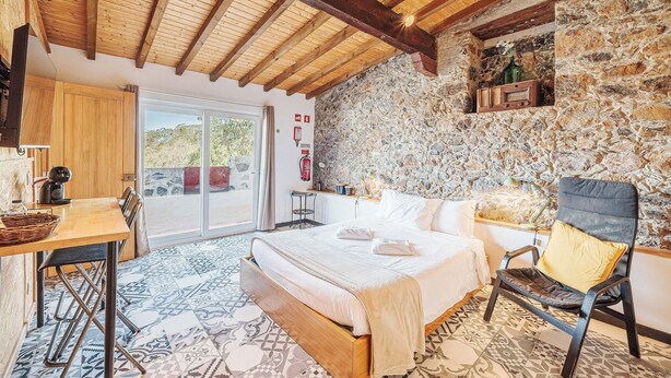 Un dormitorio con cama, una silla, una mesa, un muro de piedra y un techo de madera.