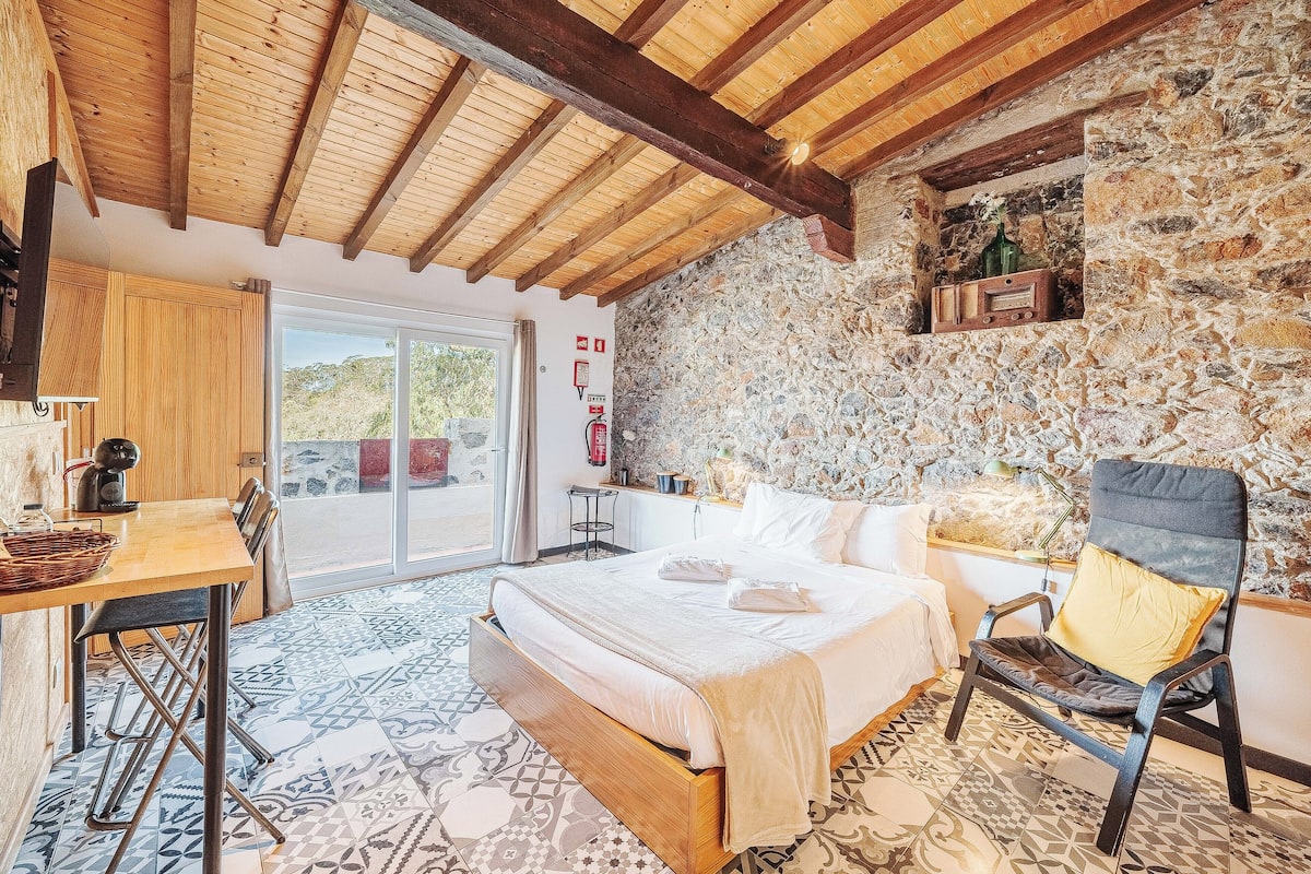 Un dormitorio con cama, una silla, una mesa, un muro de piedra y un techo de madera.