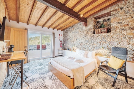 A bedroom with a bed, a chair, a table, a stone wall, and a wooden ceiling.