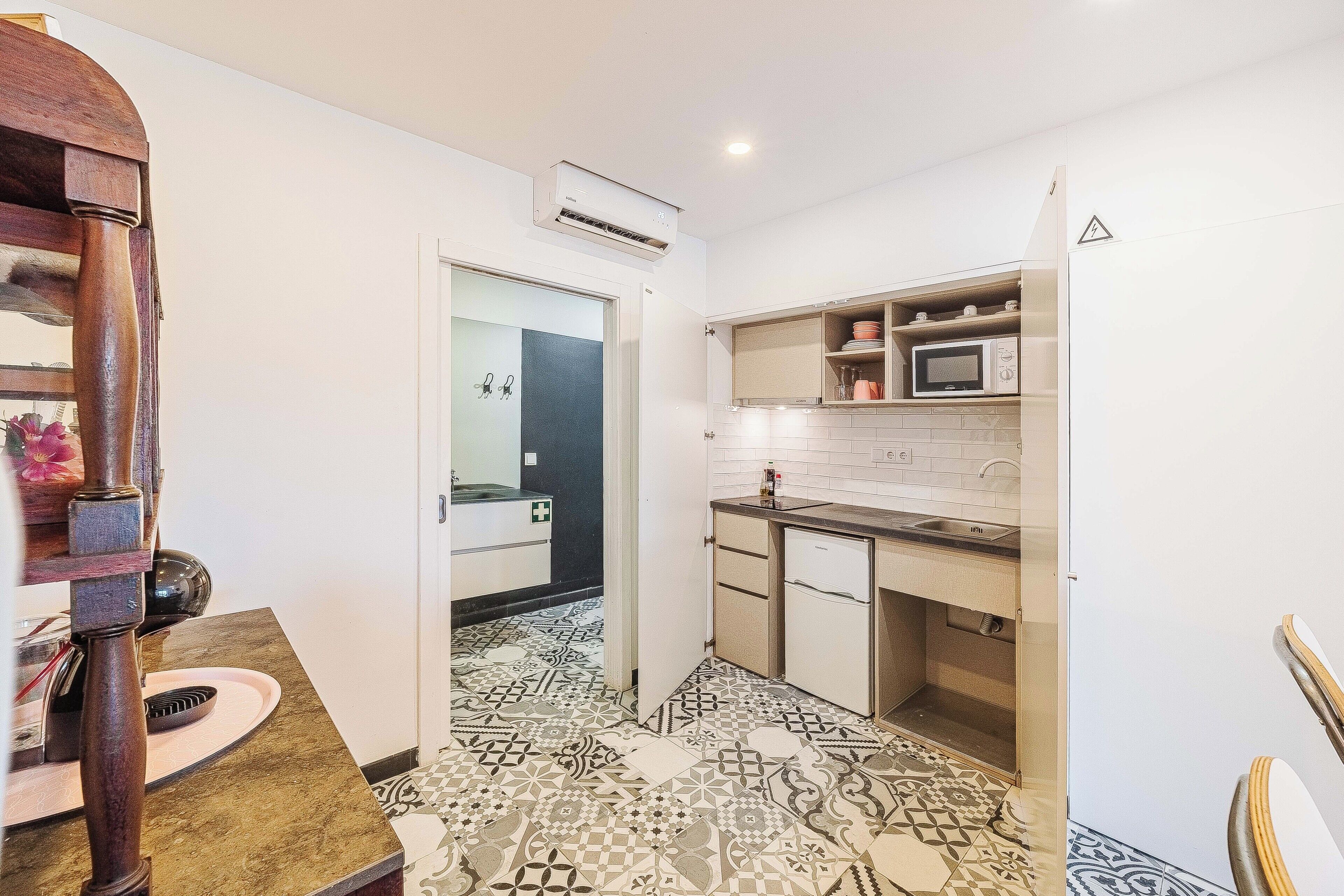 A compact kitchen with a microwave, sink, and cabinets. A patterned floor and a door leading to another room are also visible.