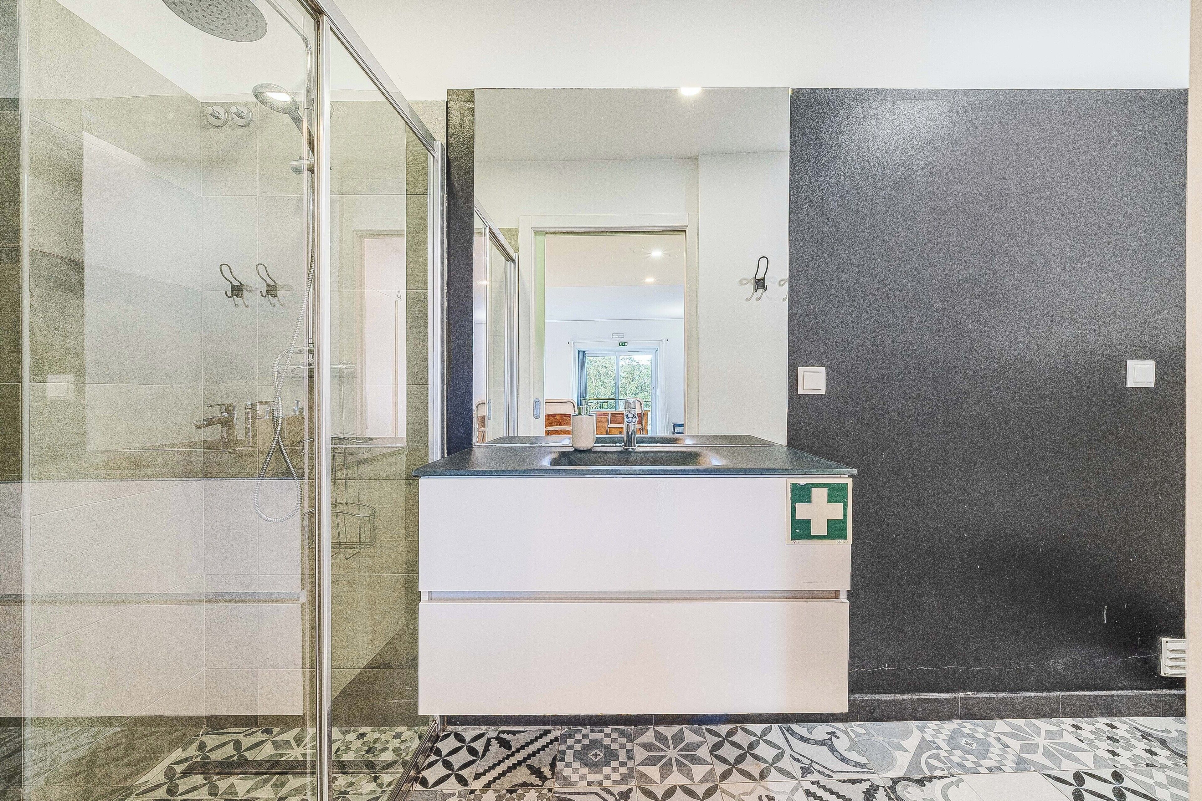 Un baño con cabina de ducha de vidrio, un lavamanos con un cartel de cruz verde y baldosas de suelo estampadas.