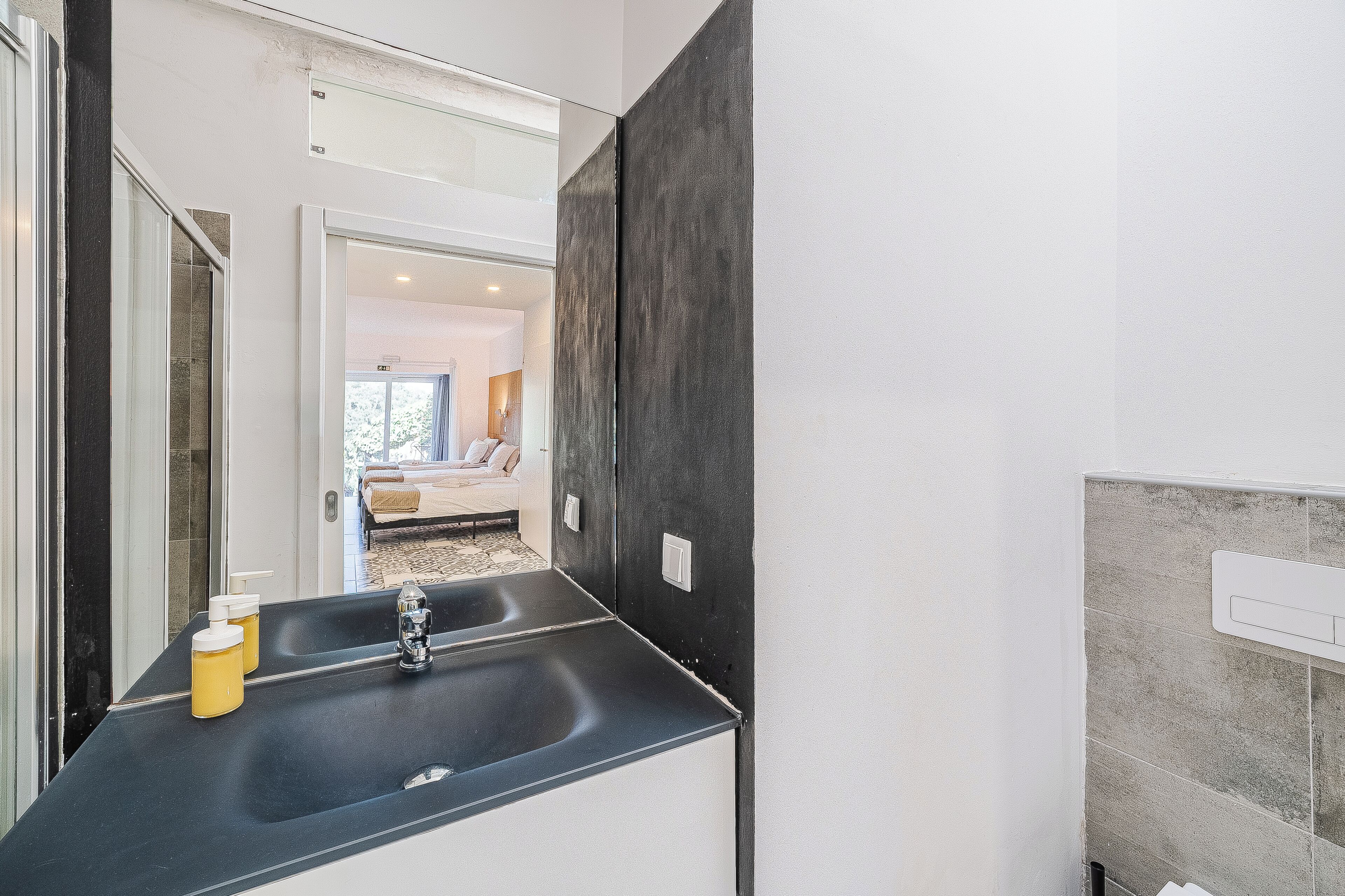 A modern bathroom with a black sink, a mirror, and a view into a bedroom with a bed and a window.