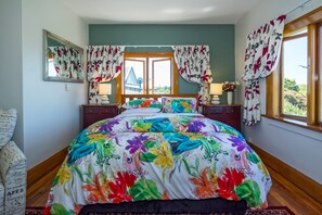 A bedroom with a floral bedspread, a wooden headboard, a window with curtains, and a view of a building outside. - The Sanctuary Beach Side (Timaru)