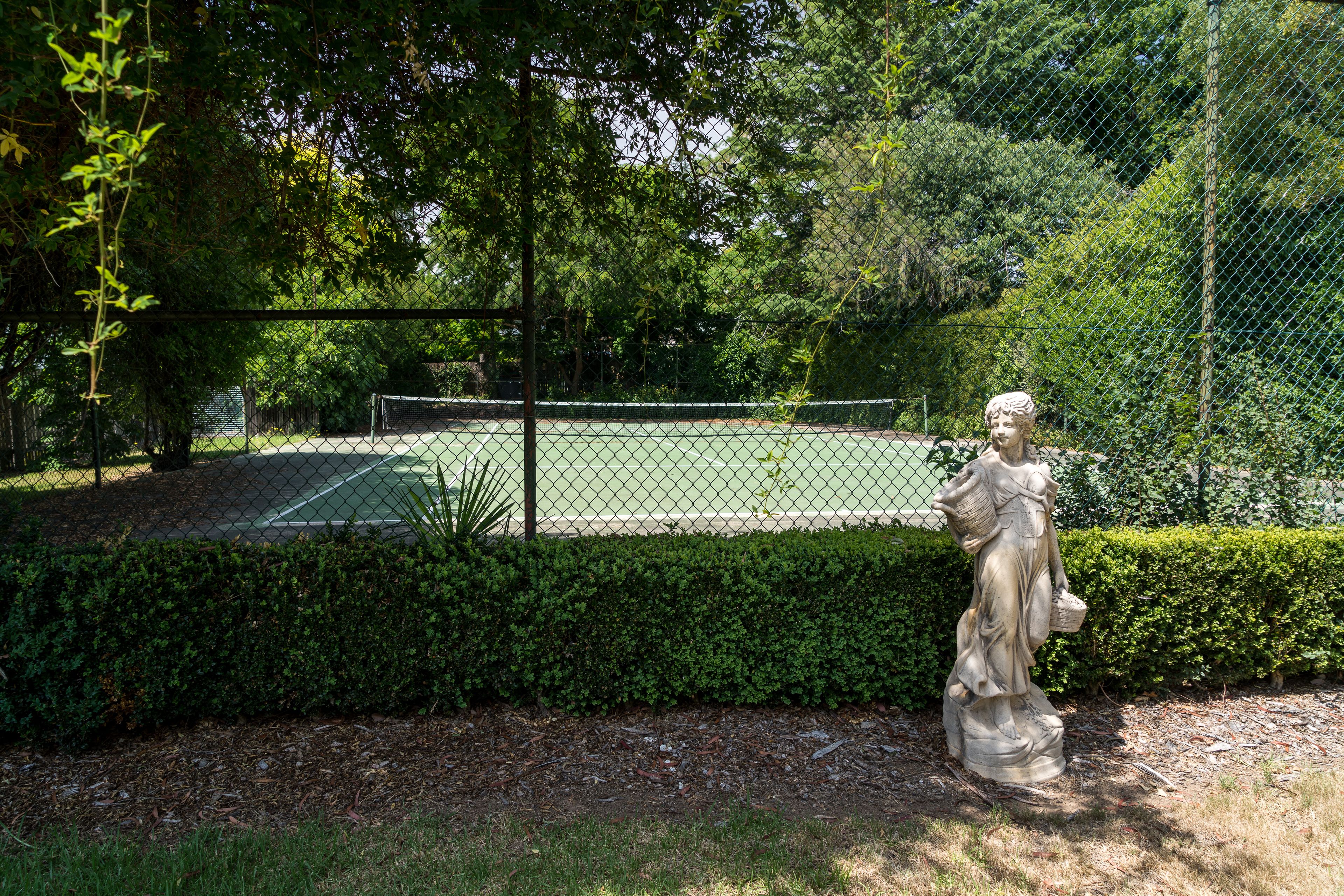 Un campo da tennis circondato da una rete metallica, con una statua di una figura che regge un cesto in primo piano.