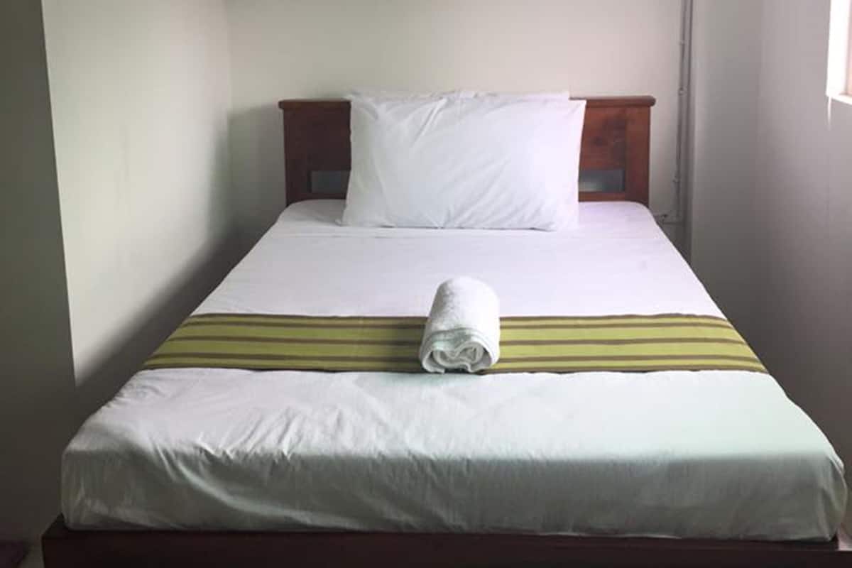 Una cama individual con cabecera de madera, ropa de cama blanca y una manta a rayas. Un toalla enrollada está sobre la cama.