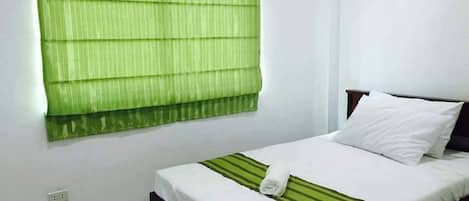 Una cama bien hecha con sábanas blancas y una colcha verde a rayas, un cabecero de madera y una persiana verde.