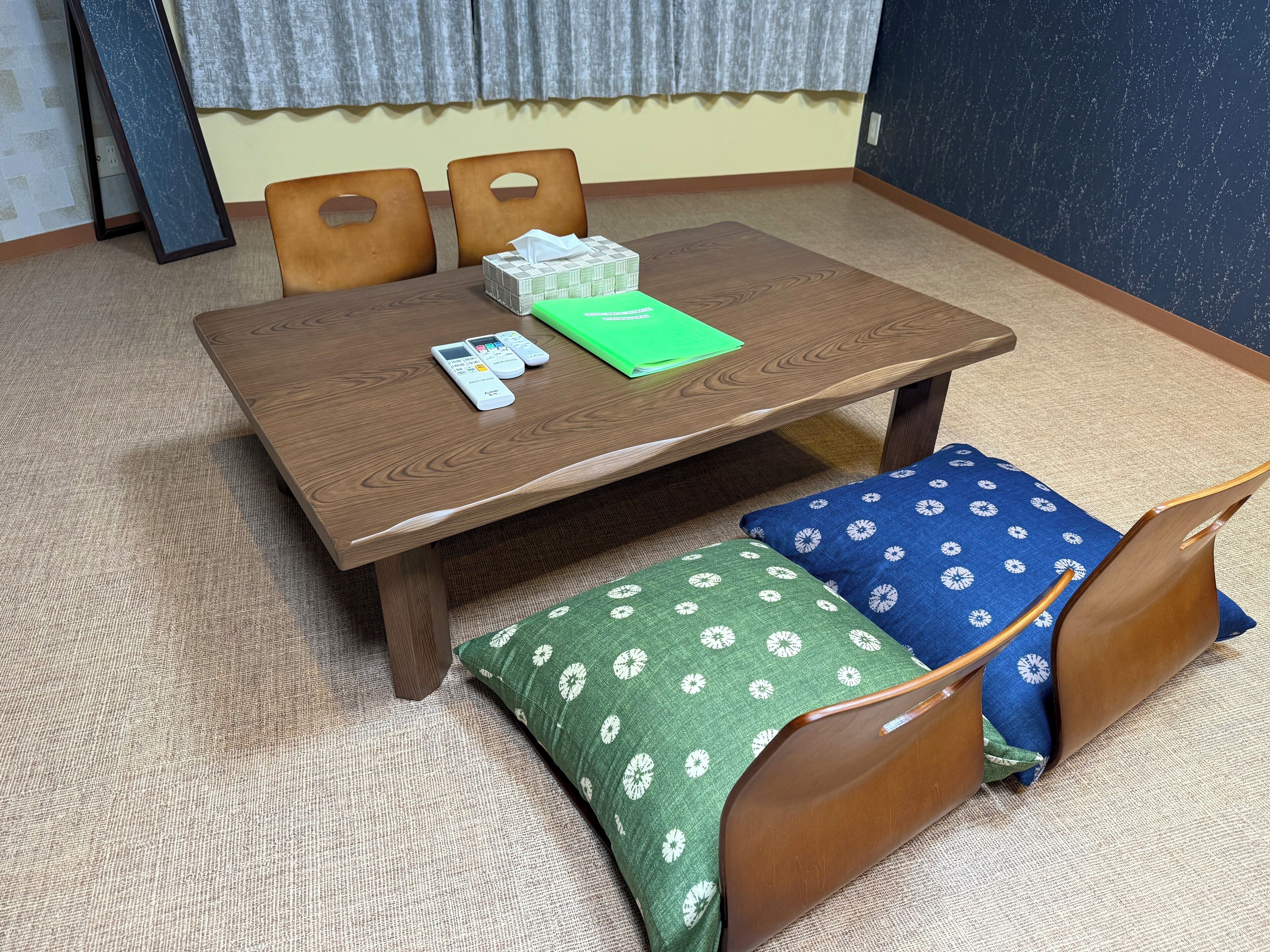 A room with a wooden table, two chairs, a green and blue patterned cushion, and a tissue box.