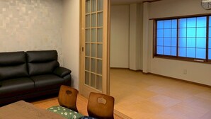 Una habitación con un sofá de cuero negro, una mesa de comedor de madera con sillas, un gran ventanal corredero y vistas a través de una ventana con puertas shoji.