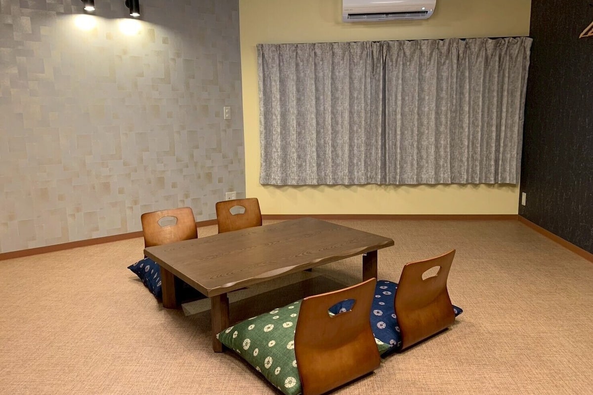 A room with a wooden table and chairs, an air conditioning unit, a patterned curtain, and a textured wall.