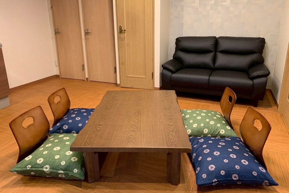 A room with a wooden table, four chairs with floral cushions, a black leather sofa, and wooden doors.