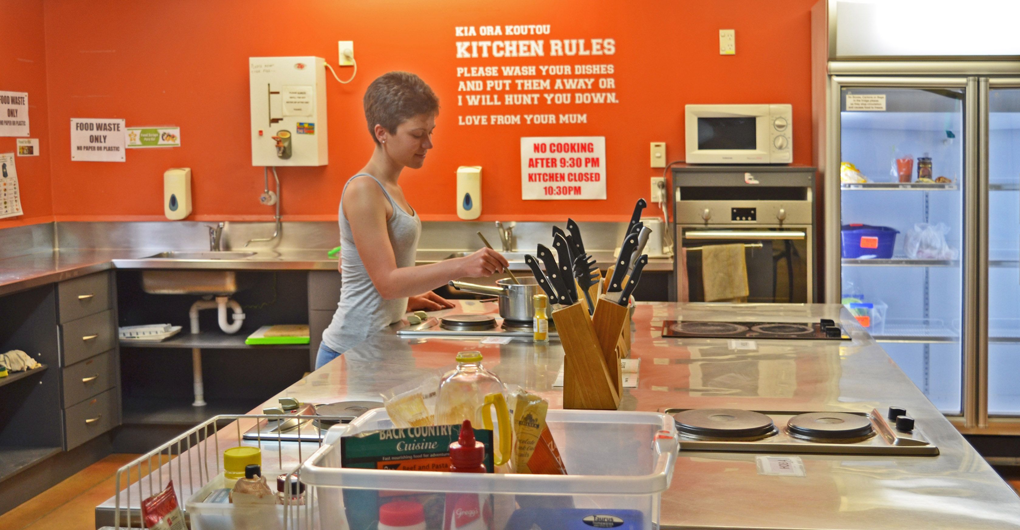A kitchen with a woman preparing food, a sign with kitchen rules, and various kitchen appliances.