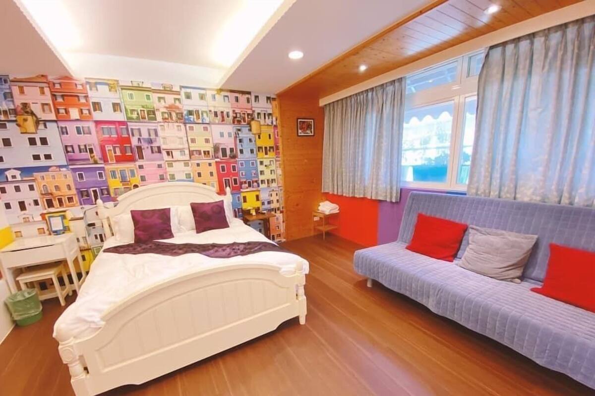 Ein Schlafzimmer mit Bett, einer Couch, einer Wand mit farbenfrohen Wandbildern und einem Fenster mit Vorhängen.