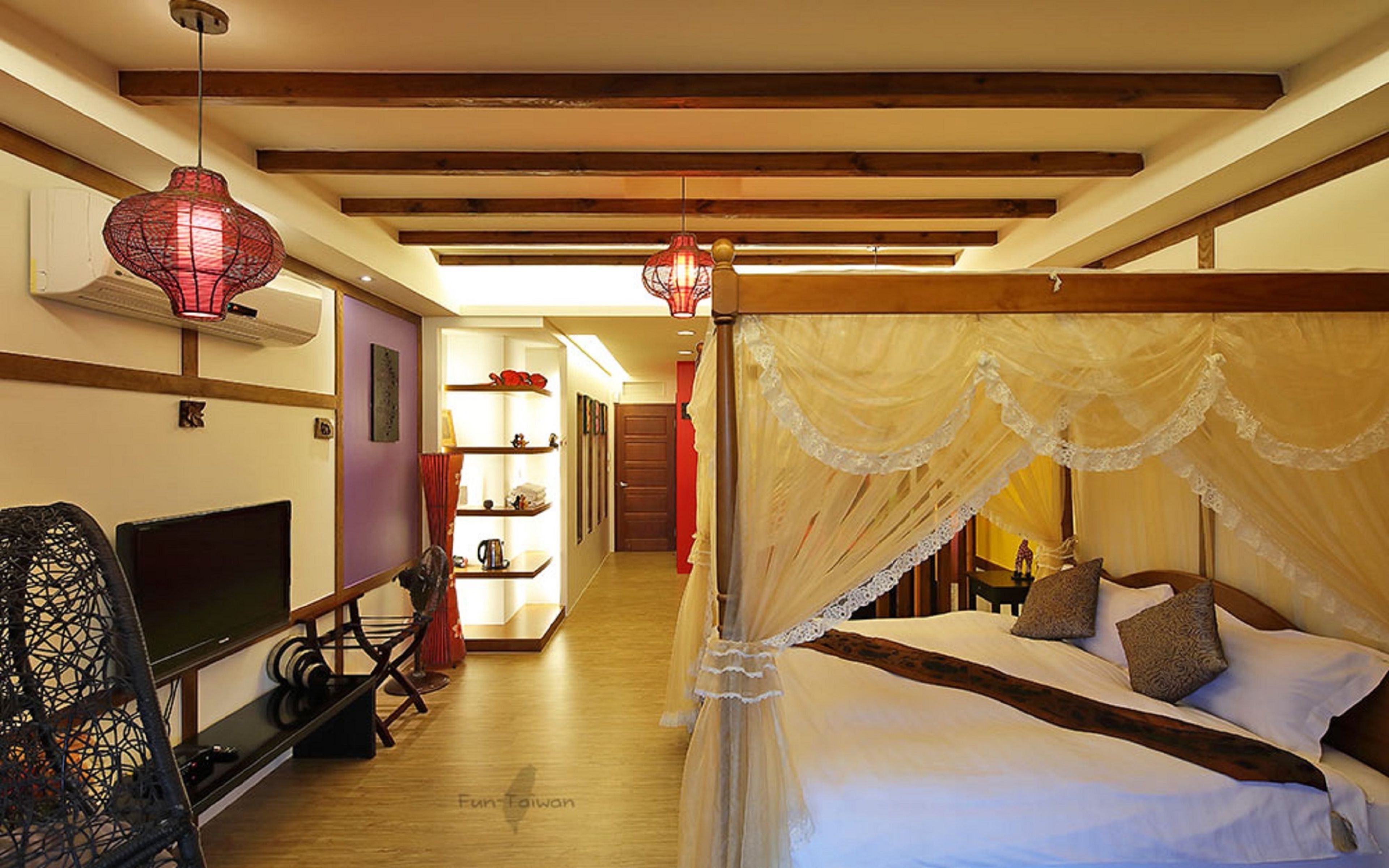 Una habitación con una cama, un televisor, una estantería con objetos y una lámpara colgante roja.