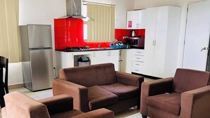 A modern kitchen with white cabinets, a red backsplash, and stainless steel appliances. There are two brown sofas in the living area. - Best View Apartments (Nadi)