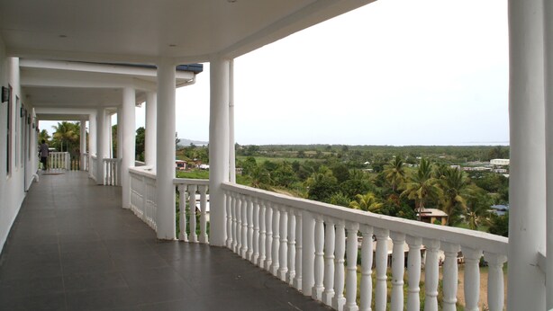 A covered corridor with white balustrades and columns, leading to a view of a green landscape with palm trees. - Best View Apartments (Nadi)