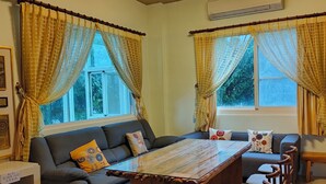 Una sala moderna con un sofá, reposapiés y una mesa de centro de madera. Hay cortinas en las ventanas y un equipo de aire acondicionado montado en la pared.