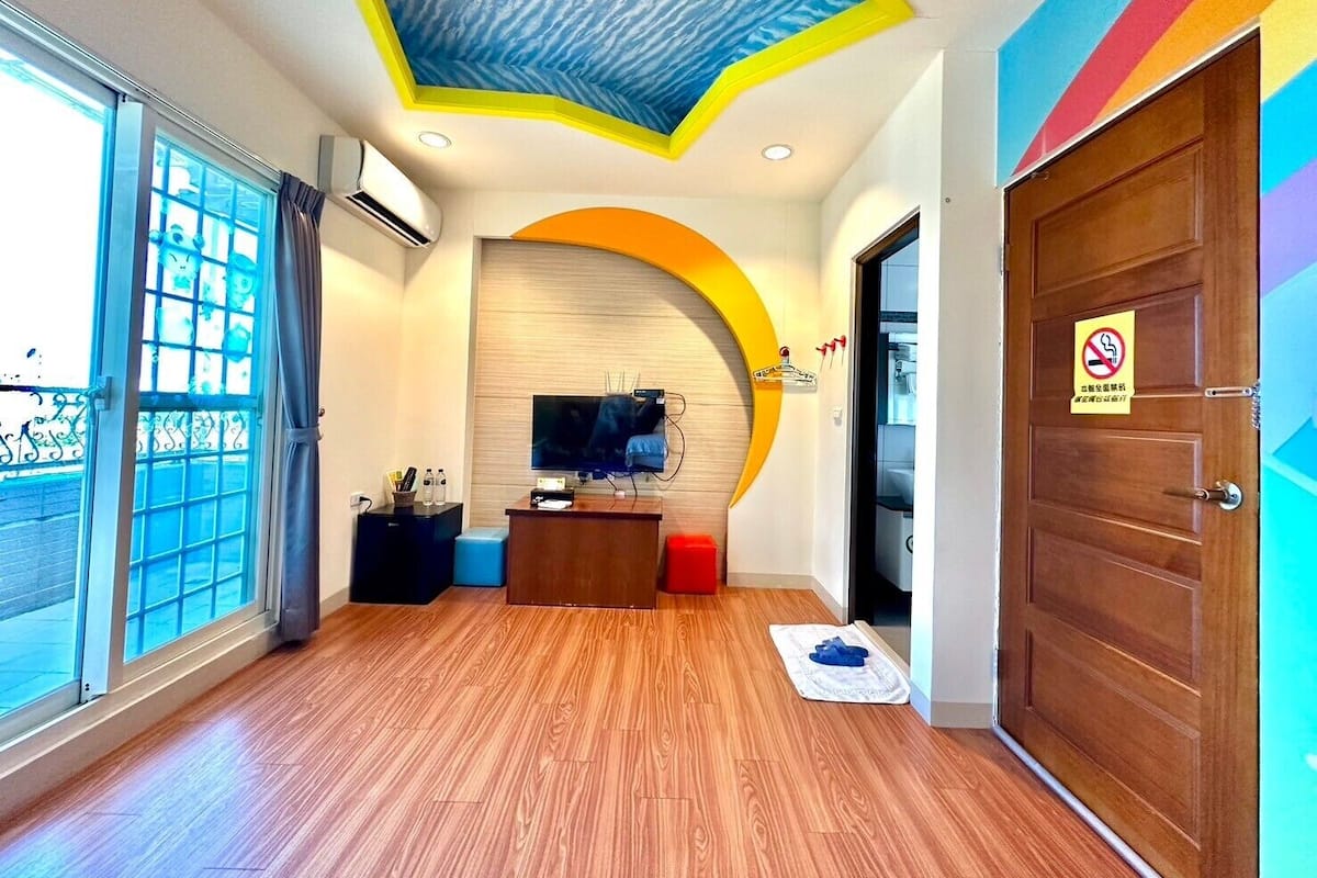 A modern hotel lobby with a wooden floor, a reception desk, a flat-screen TV, and a large window.