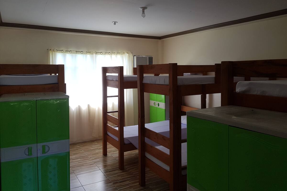A dormitory room with bunk beds, a green wardrobe, and a window with curtains.