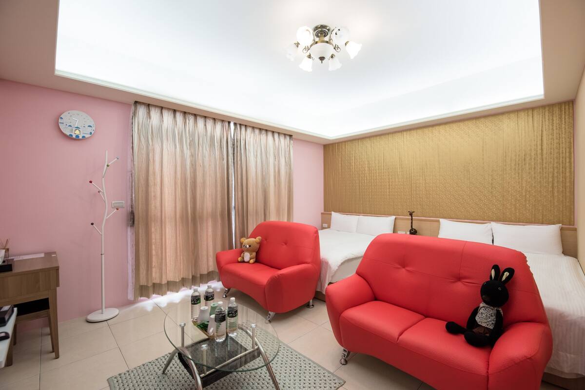 Habitación de hotel con una cama, dos sofás rojos, una mesa de centro de vidrio, un escritorio, una lámpara de pie, un osito de peluche y un reloj en la pared.