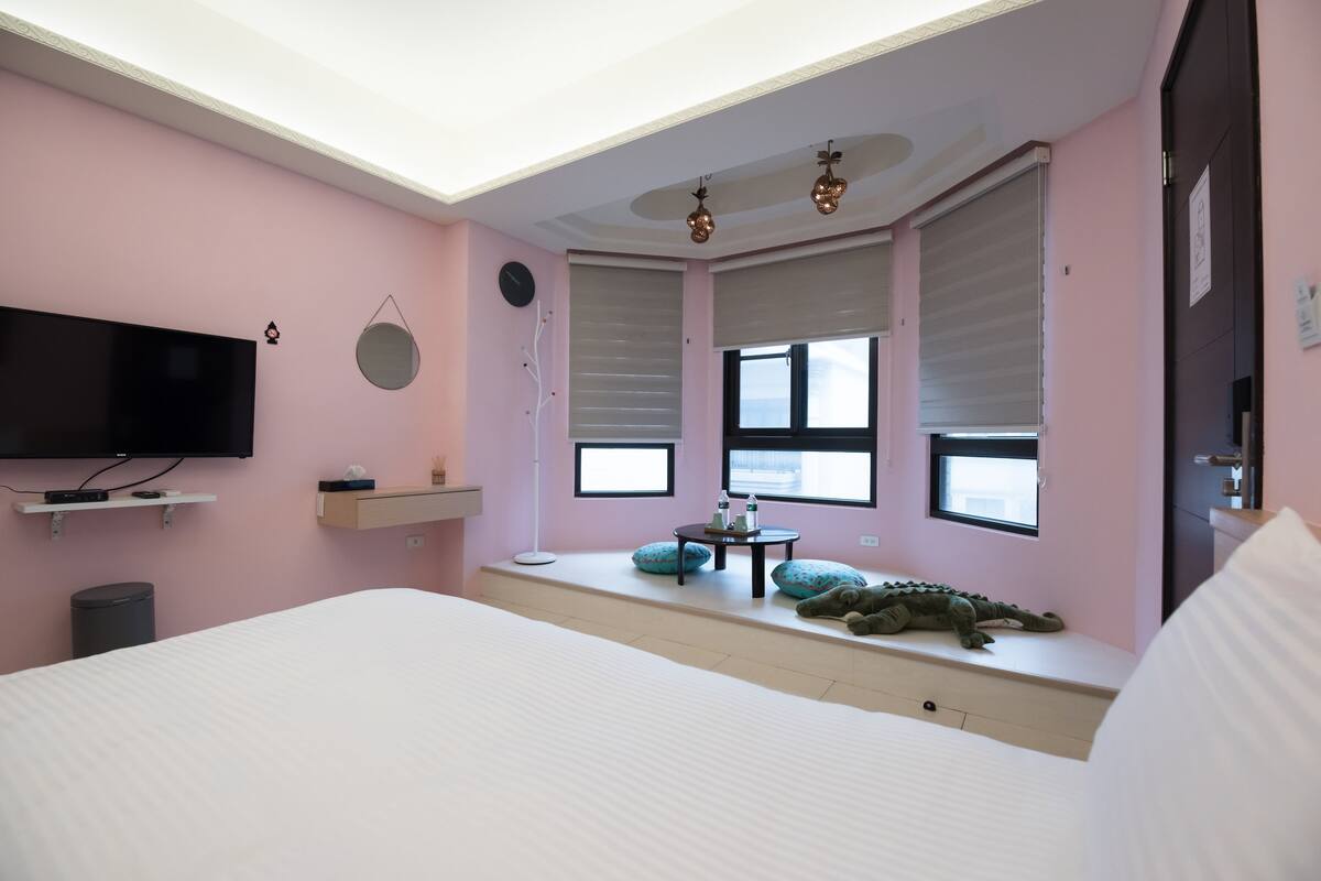 Un dormitorio con una cama grande, un televisor instalado en la pared, una mesita con una lámpara y una ventana con vistas.