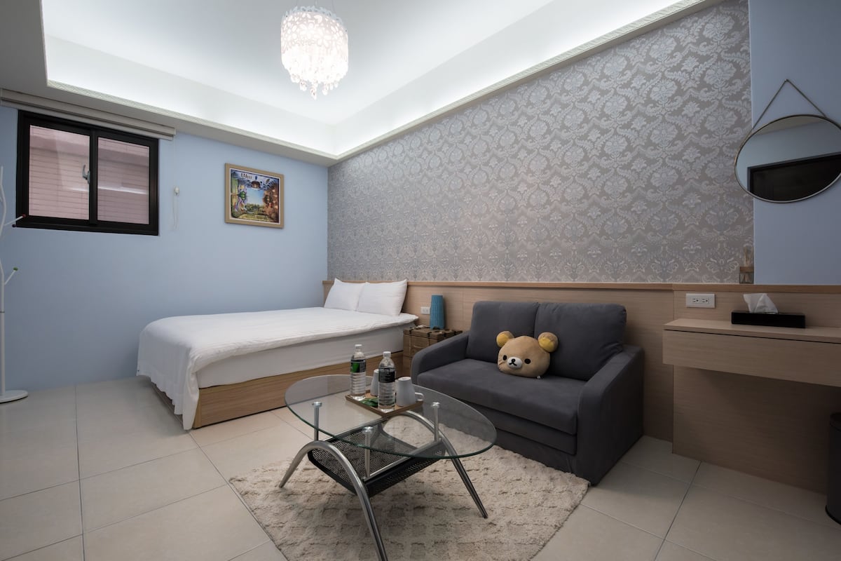 Un dormitorio compacto con cama, una mesita de centro de vidrio, un sofá con un oso de peluche y una zona de escritorio.