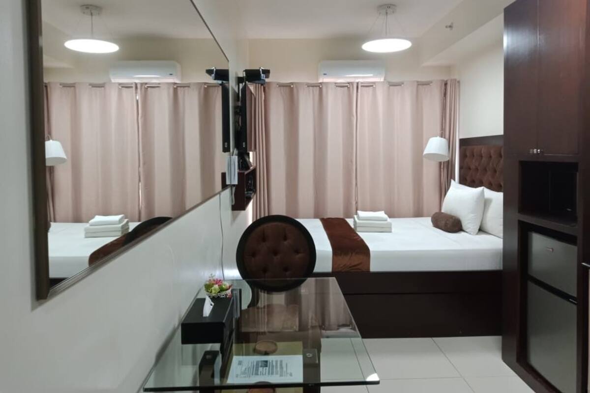 A modern hotel room with a glass table, a bed with a brown headboard, and a large mirror reflecting the room.