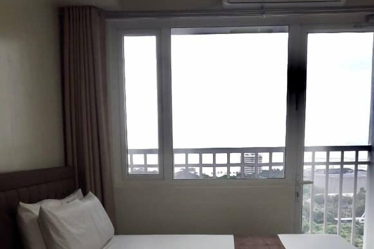 A single bed with a headboard, a bedside table, and a window with a view of buildings and greenery.