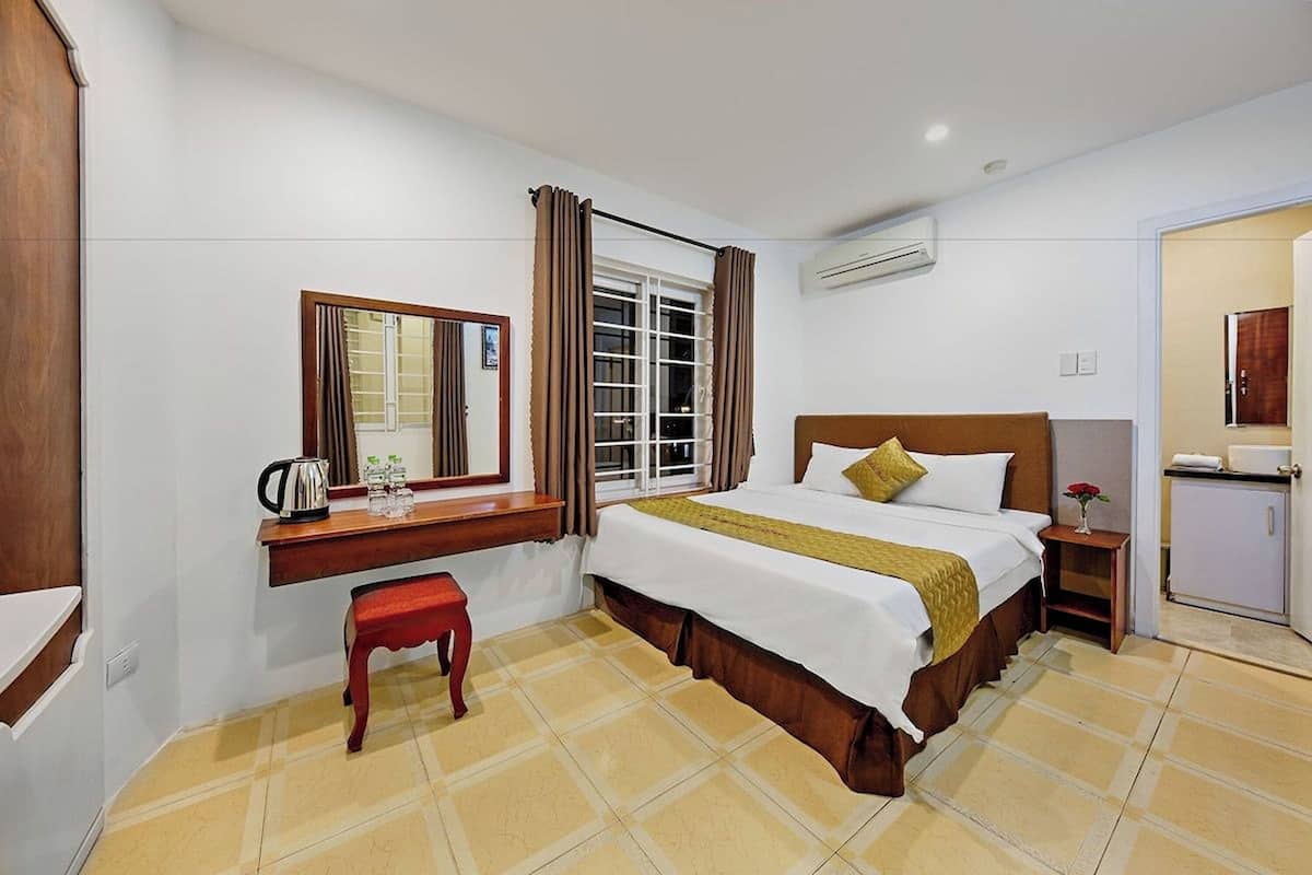 A hotel room with a bed, a desk with a mirror, a window with curtains, a small table with a vase, and a cabinet.