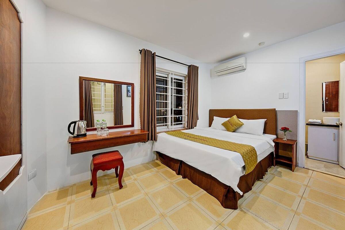 Quarto de hotel com uma cama, uma pequena escrivaninha com chaleira, um espelho e uma janela com cortinas.