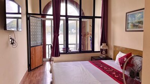 A hotel room with a large bed, a nightstand, a lamp, a painting, and a view of a body of water through large windows. - Le Pont Bungalow Hostel (Hai Phong)
