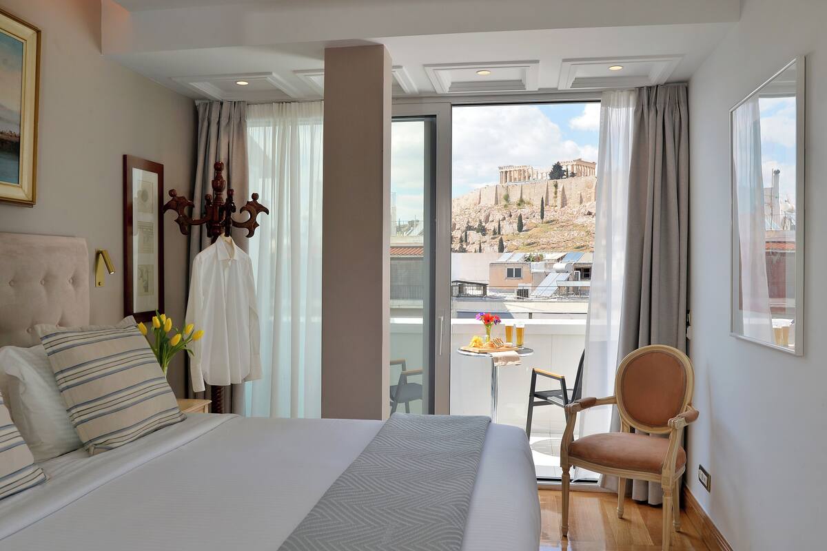 A hotel room with a bed, a chair, a table with a drink and fruit, and a view of a historic building.