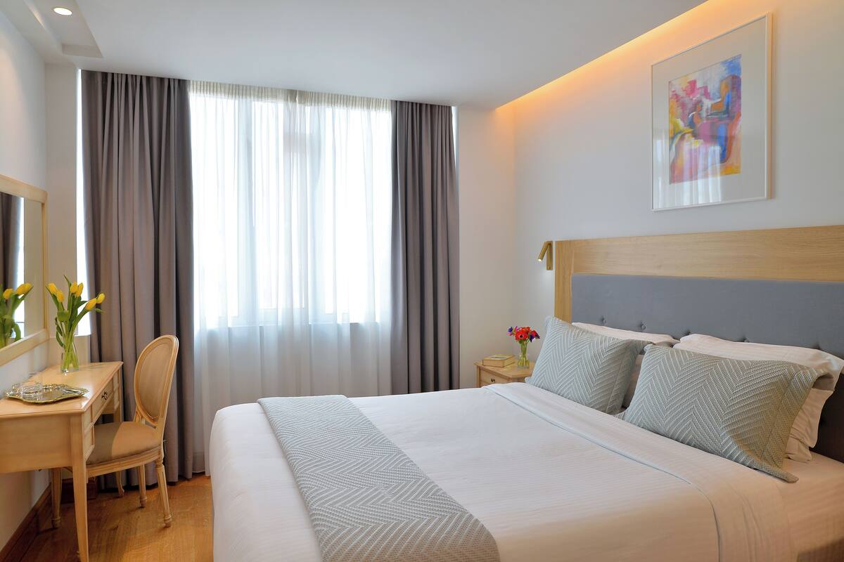A hotel room with a large bed, a desk with a chair, a nightstand with a vase of flowers, and a painting on the wall.