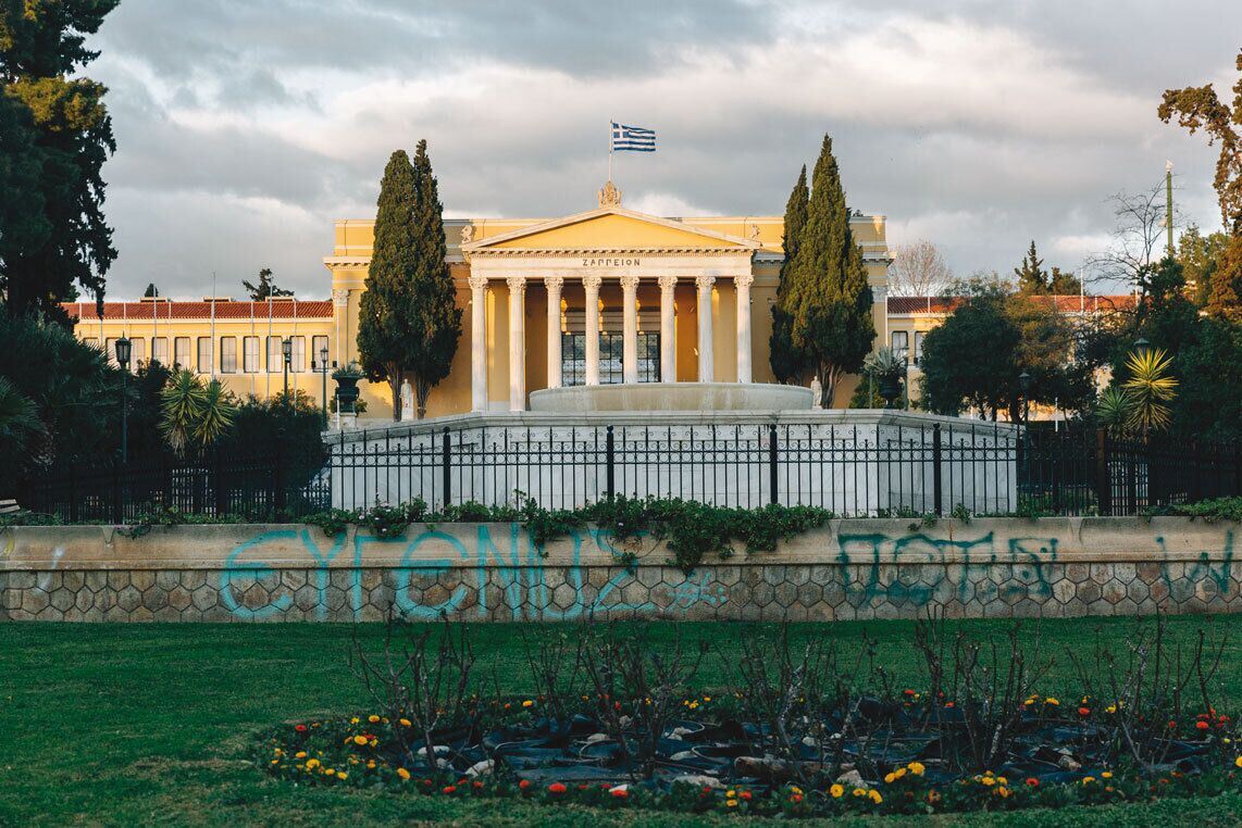 A classical building with columns, a flag, and a fountain in front.