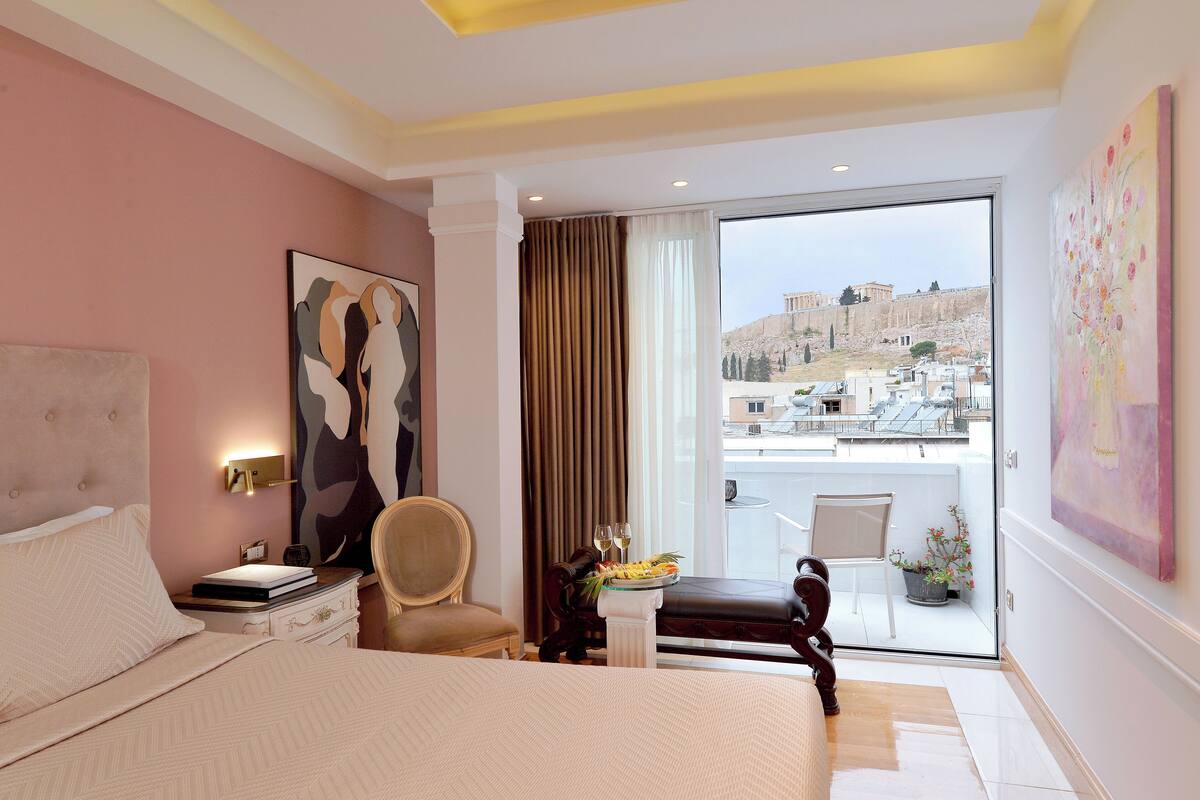 A hotel room with a bed, a chair, a small table with a fruit basket and wine glasses, and a balcony with a view of ancient ruins.