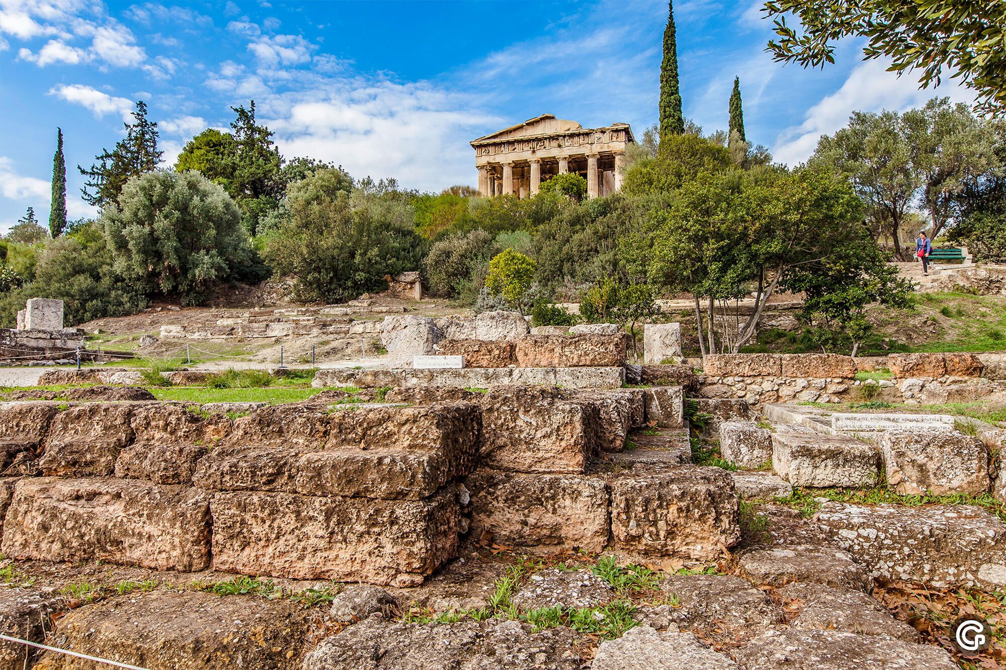 Ancient ruins with columns and stone blocks, surrounded by trees and a clear sky.