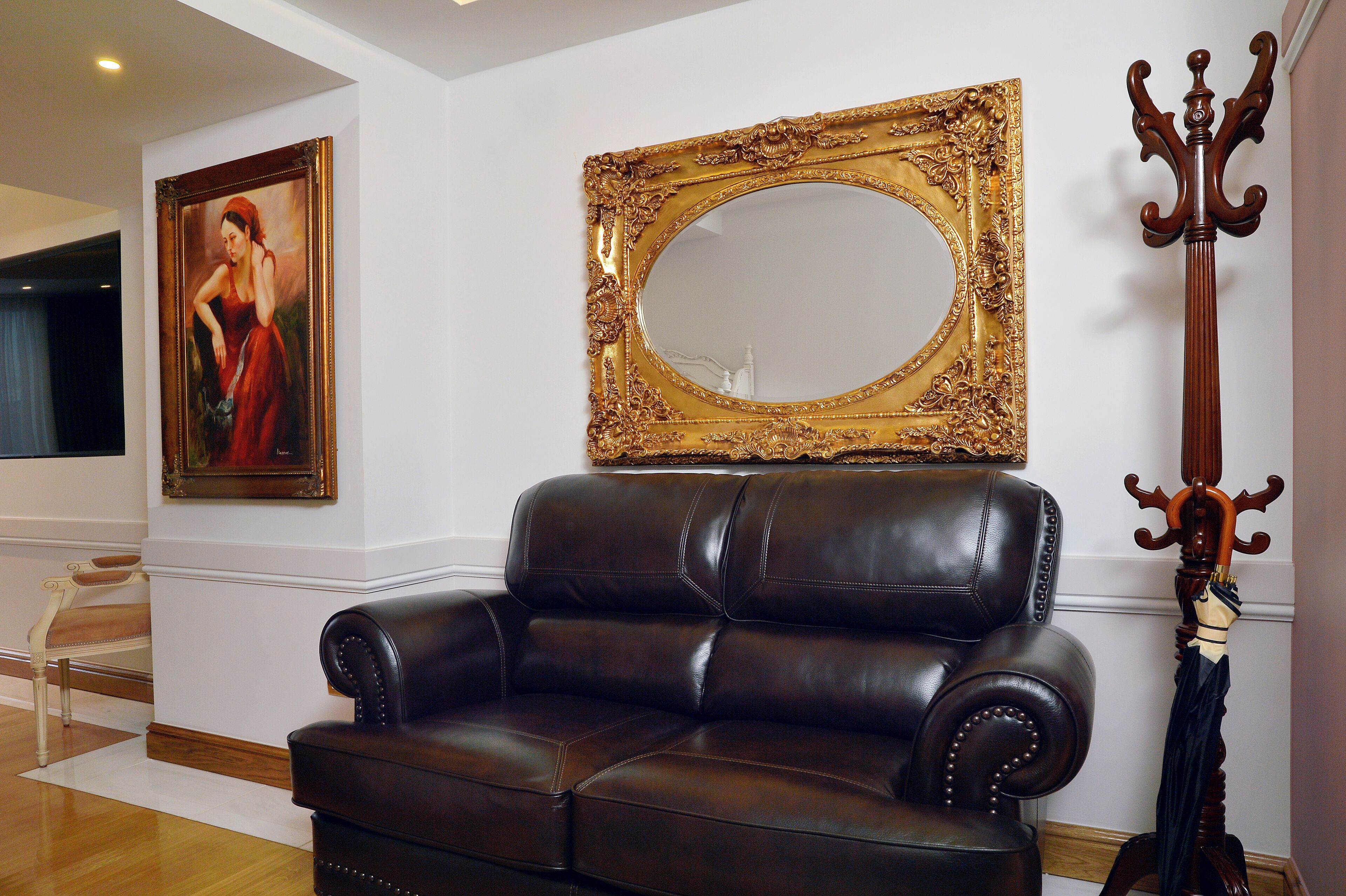 A dark brown leather sofa, a large ornate mirror, and a wooden coat stand in a room with white walls and a wooden floor.