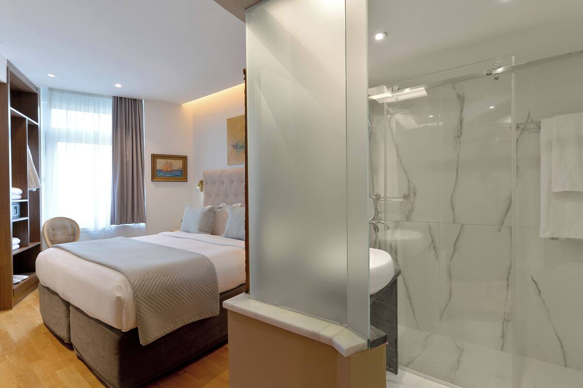 A hotel room with a bed, a bathroom with a glass shower enclosure, and a view of the sea through the window.