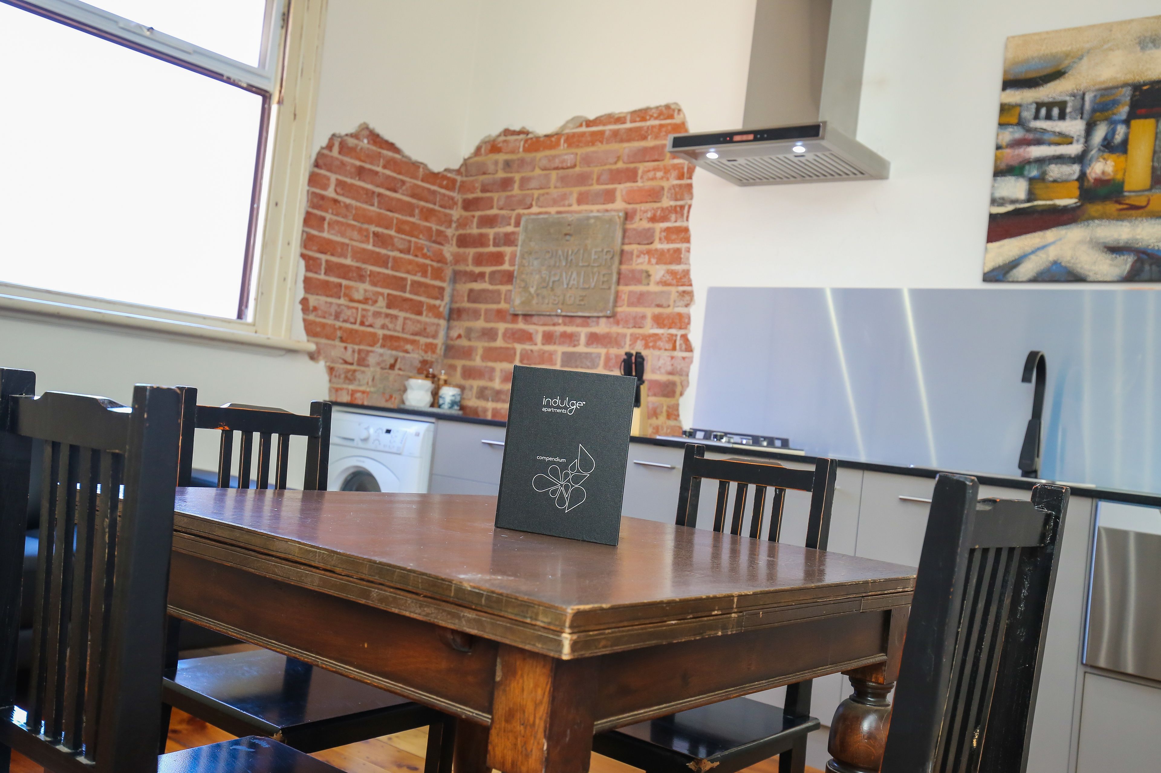 A dining area with a brick wall, a table with chairs, a kitchen in the background, and a painting on the wall.