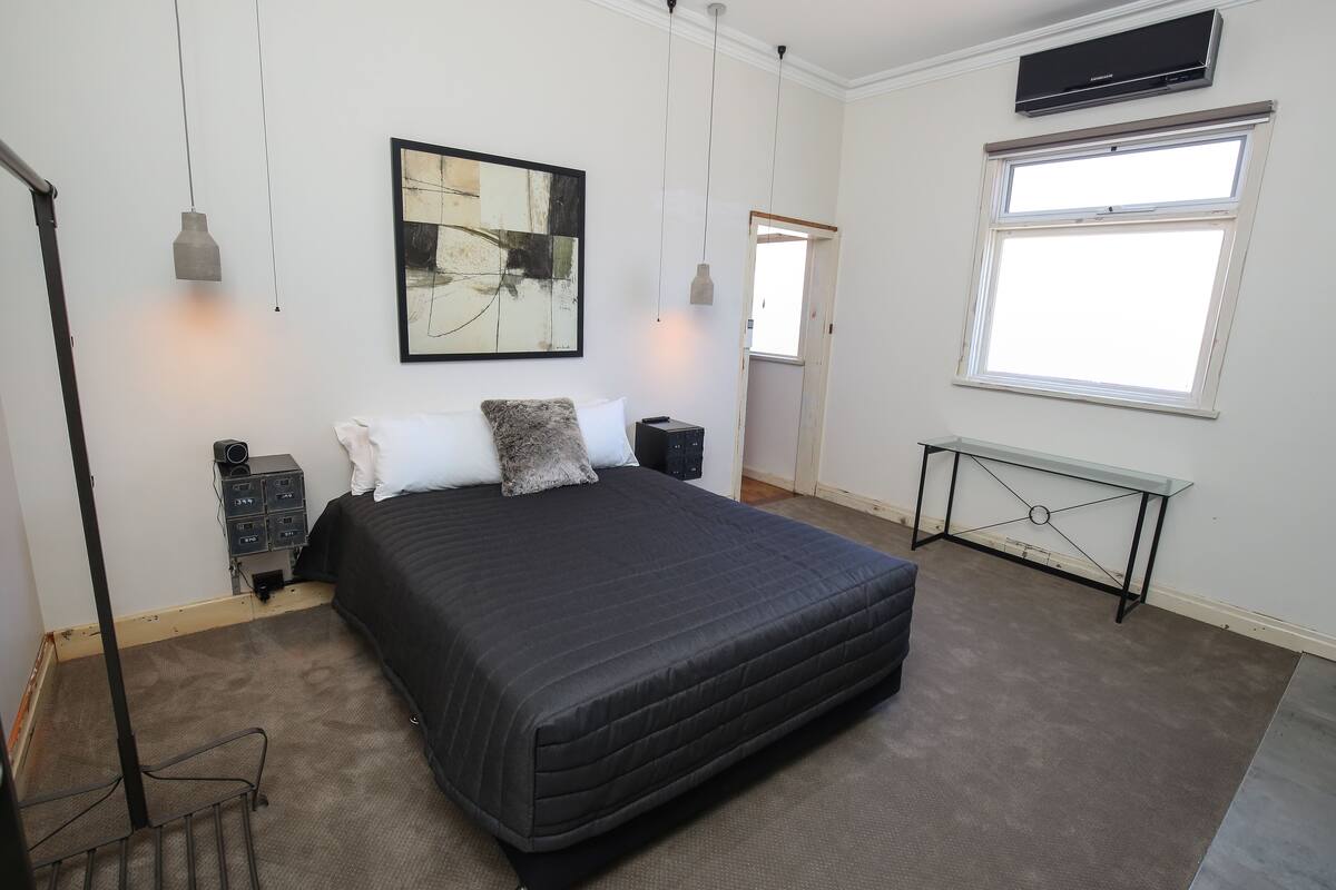 A bedroom with a large bed, a desk, a chair, a standing lamp, a radiator, and a window.