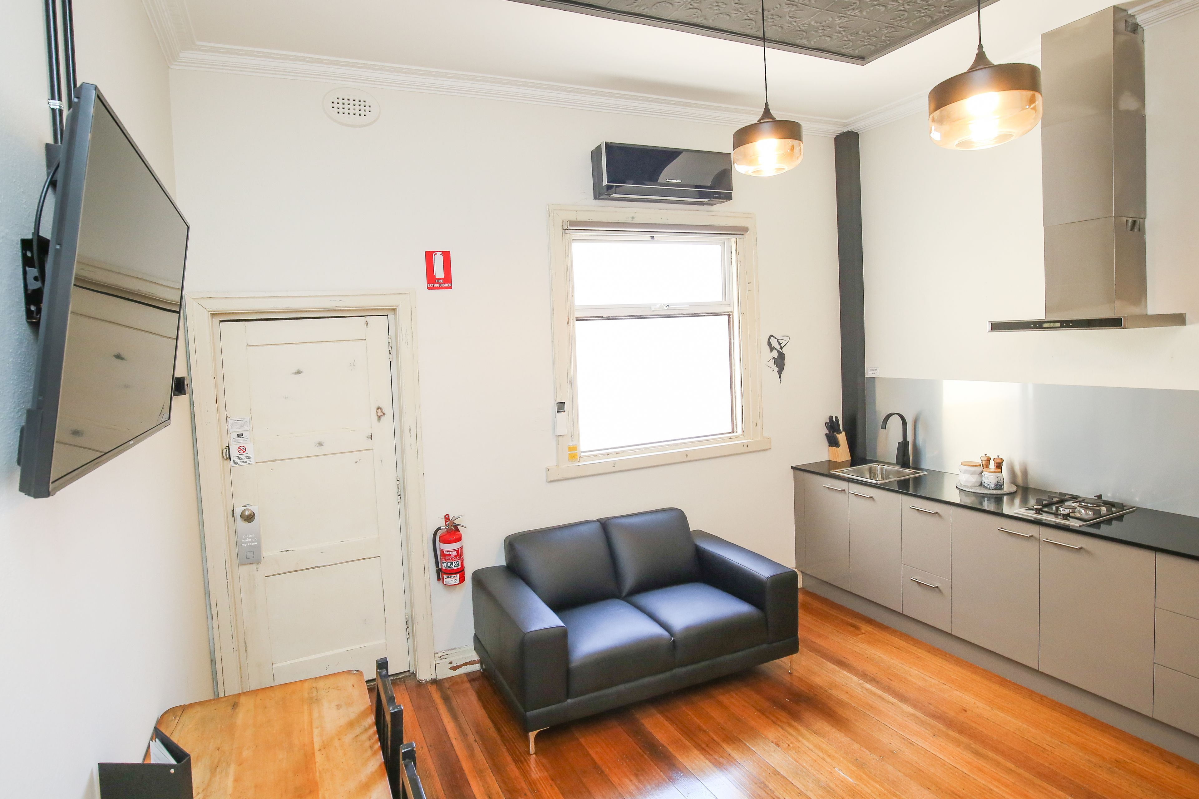 A compact living space with a kitchenette, a black leather sofa, wooden flooring, and a window providing natural light.